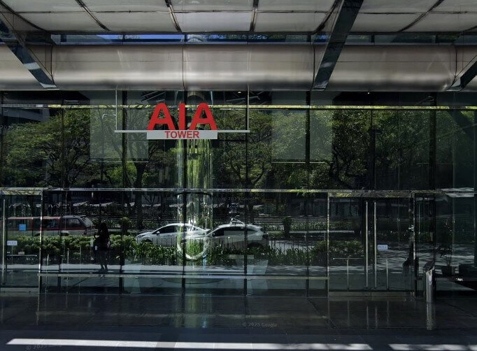 A ground-level view of the AIA Tower entrance, showing glass walls reflecting trees and a street with parked cars.