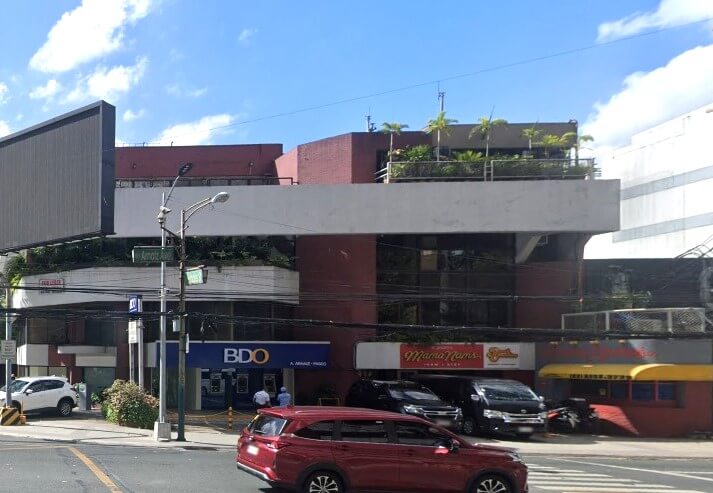 The exterior of the BDO A. Arnaiz-Paseo Branch building. The bank entrance is visible at street level, flanked by signs for the Mama Nams restaurant. A red SUV passes in the foreground. The building is a two-story structure with reddish-brown brick and white trim, with some greenery on the roof