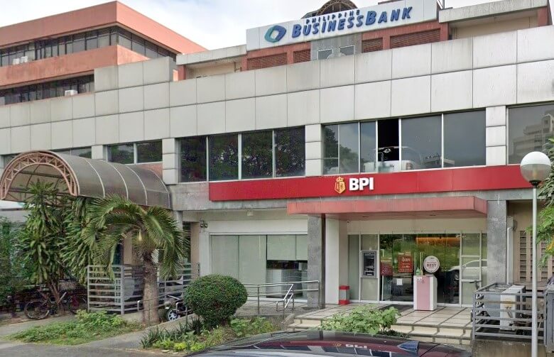 he exterior of a modern two-story commercial building. The ground floor features a BPI (Bank of the Philippine Islands) branch with a red facade and logo above the entrance, flanked by large glass windows. The second floor features a Philippine Business Bank sign