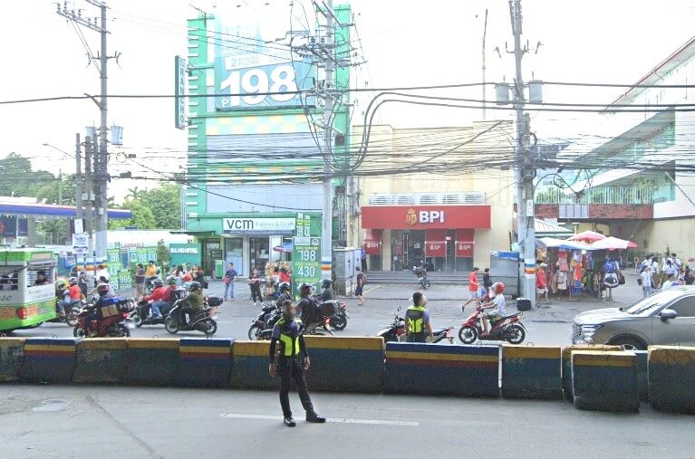 A street view of the BPI (Bank of the Philippine Islands) Muntinlupa Alabang Branch, a red building with white lettering, located on a busy road. The foreground shows a concrete barrier and a security guard in uniform facing away from the camera