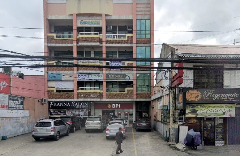 A street-level photo of a multi-story commercial building housing various businesses, including a BPI bank branch at the ground level, with a parking area in front containing several cars
