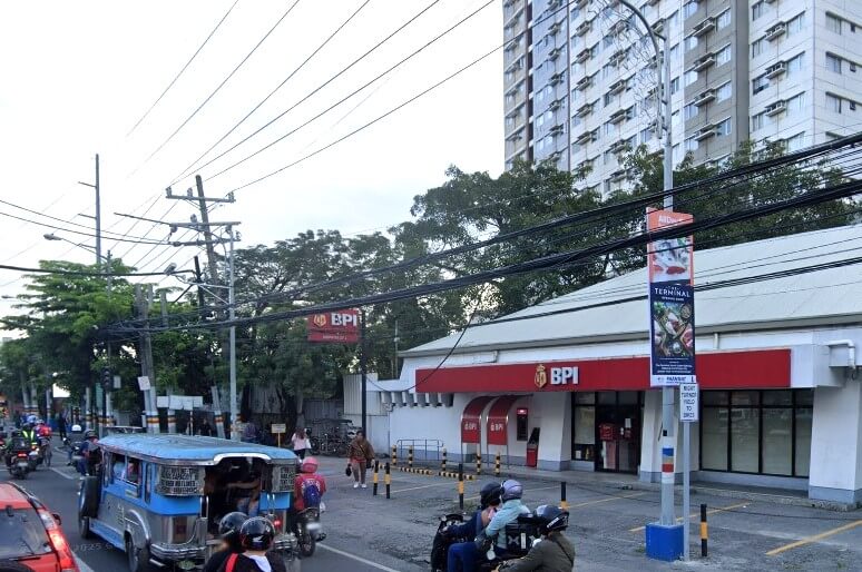 BPI Muntinlupa South Park Branch. The building has white walls and a red awning over the main entrance and ATM. A high-rise residential building is visible behind the bank on the right