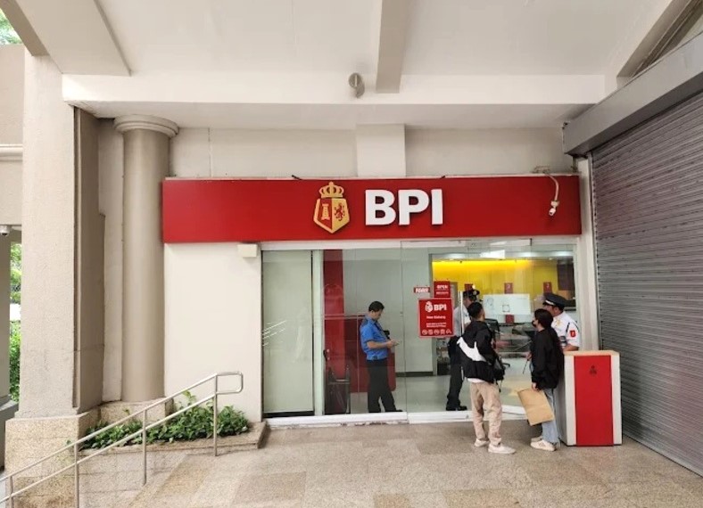 Exterior view of a BPI (Bank of the Philippine Islands) branch entrance, featuring the bank's red and gold signage. Several people are standing near the glass doors, including a security guard in uniform