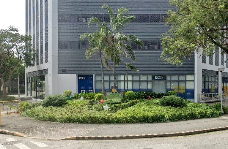 The exterior of a modern commercial building with a BDO bank branch office visible on the ground floor, set behind a lush garden and sidewalk.