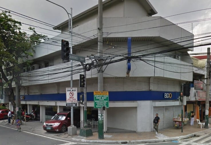Exterior view of a BDO bank branch building at a street corner, with utility poles, traffic lights, and a red car parked nearby.