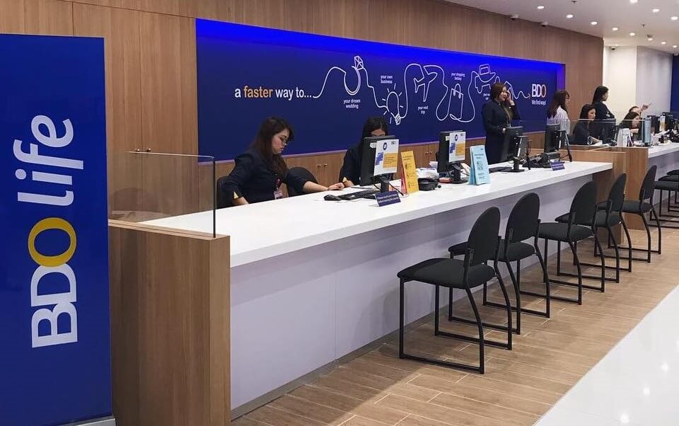 Indoor view of the BDO A. Bonifacio Ave. - Cloverleaf bank branch, showing the long service counter where several female staff are assisting customers. The counter has a white top and a blue wall behind it with the BDO logo and the text "a faster way to…" along with simple line art drawings. A large "BDO life" sign is visible on the left