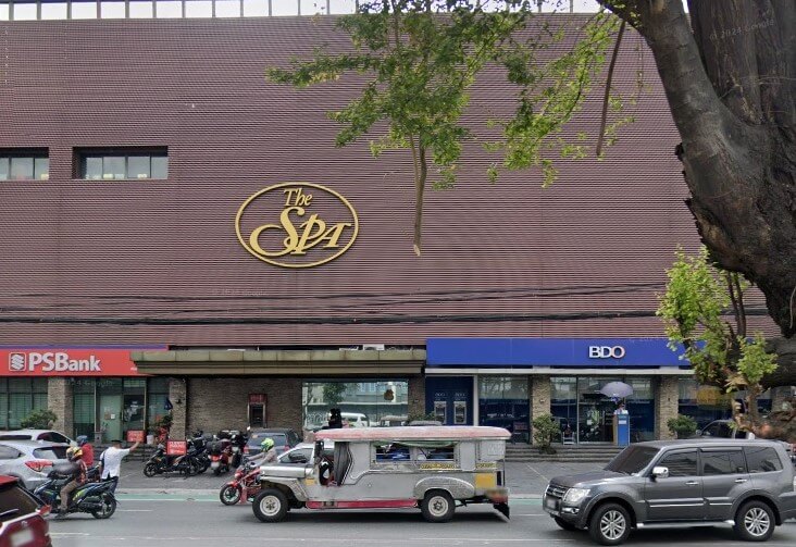 Street-level view of a commercial building featuring a large gold sign for "The Spa" on the upper floor, with a BDO bank branch and a PSBank branch at the ground level. A jeepney and several other vehicles are visible on the street in the foreground
