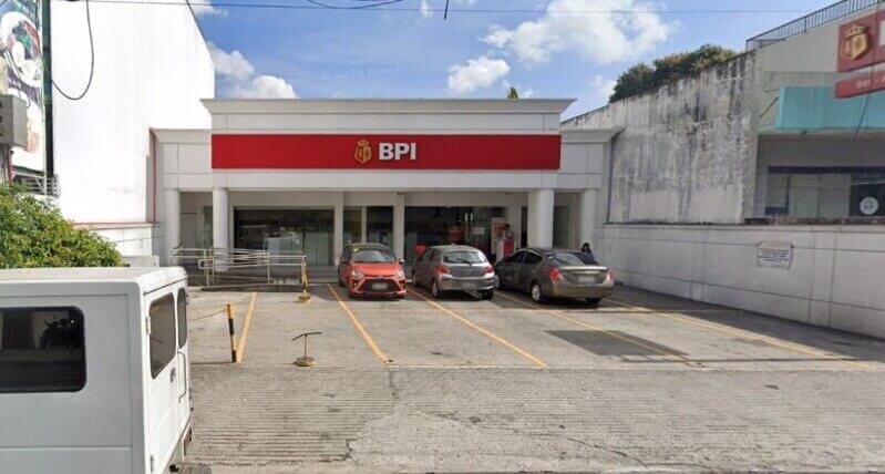 A street-level view of the BPI Bel Air Paseo branch building, featuring a white facade with a prominent red horizontal sign displaying the BPI logo. Three cars are parked in the paved lot in front of the entrance, and a white van is partially visible in the foreground