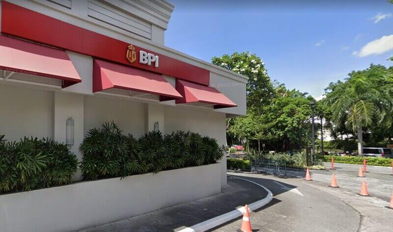 Exterior view of a BPI (Bank of the Philippine Islands) branch featuring a white building with a prominent red sign and awnings, situated next to a paved driveway with orange traffic cones and lush green trees in the background