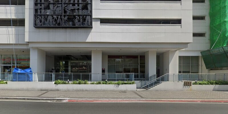 Exterior view of a BPI branch located at the ground floor of a modern white building, featuring large glass windows, concrete pillars, and a grey metal railing along the sidewalk