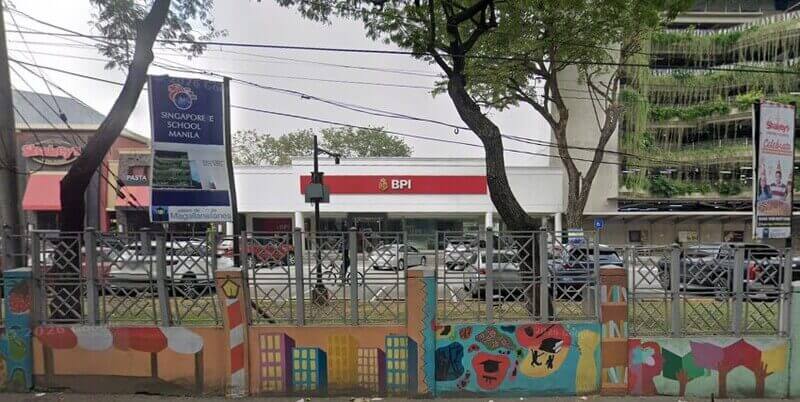 A street-level view of a BPI (Bank of the Philippine Islands) branch in Magallanes, Manila, situated between a Shakey’s Pizza parlor and a multi-level parking structure with hanging plants. In the foreground, a gray metal fence features colorful murals and a banner for Singapore School Manila