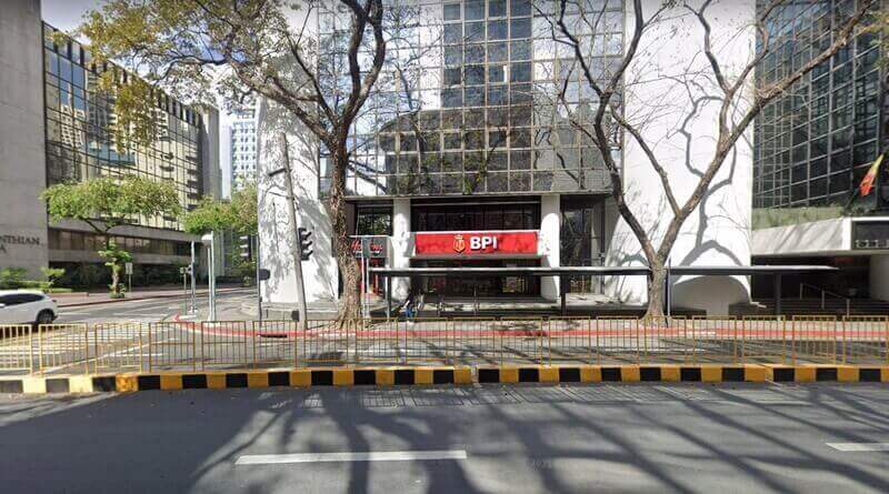 Street-level view of a BPI bank branch at the base of a modern glass-windowed building on a city corner, framed by tall trees and a yellow-and-black safety barrier in the foreground