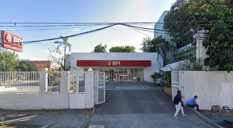 Exterior view of a white, single-story BPI (Bank of the Philippine Islands) branch featuring a prominent red sign and a gated driveway entrance