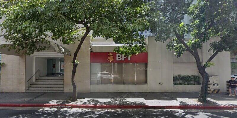 Exterior view of a BPI (Bank of the Philippine Islands) branch featuring a red sign with the gold shield logo above large glass windows, situated on a tree-lined sidewalk next to a building entrance with stairs