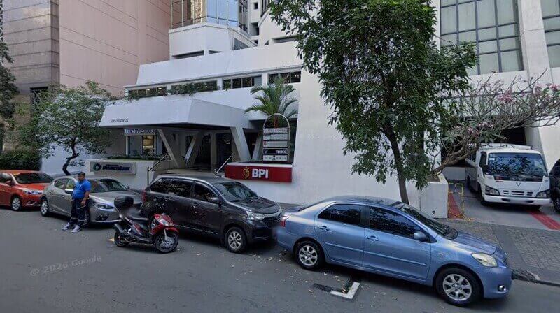 Street-level view of the BPI Alfaro branch exterior, featuring a white modern building with a red BPI sign, several parked cars and a motorcycle in front, and a white van to the right