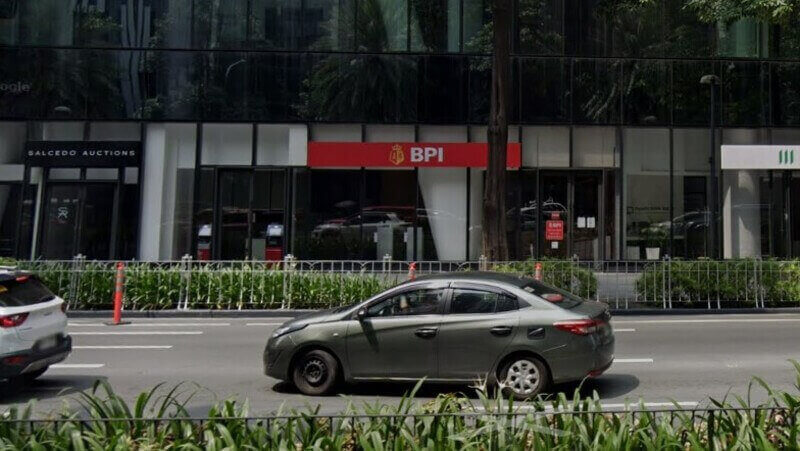 Street-level view of the Ayala North Exchange (Nex Tower) in Makati City, featuring the glass facade of the building with BPI and Manulife storefronts at the ground floor, fronted by a busy city road with passing vehicles and lush tropical greenery
