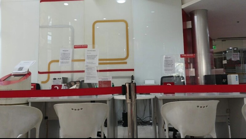Interior of a BPI bank branch featuring service desks with white chairs, protective plexiglass shields, and red queue barriers. The background shows a white wall with the bank's signature red and gold line graphics and posted service notices