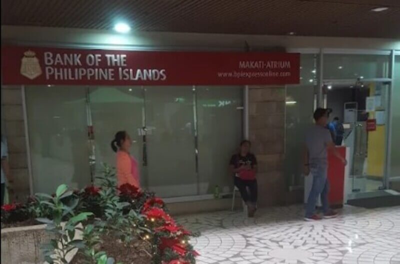 The storefront of a Bank of the Philippine Islands (BPI) branch at the Makati Atrium, featuring the red corporate signage with the bank's name and website. Several people are standing or sitting in the tiled mall area in front of the glass entrance