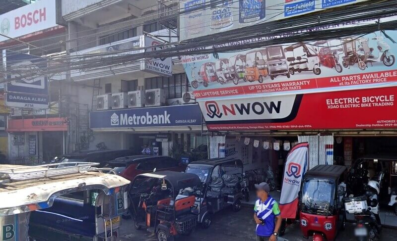 A street-level view of the Metrobank Albay-Daraga Branch in the Philippines, located next to an NWOW Electric Bicycle dealership. The scene shows several electric tricycles parked in front of the shops, various commercial signs for Bosch and paint brands, and a person in a blue shirt standing on the sidewalk