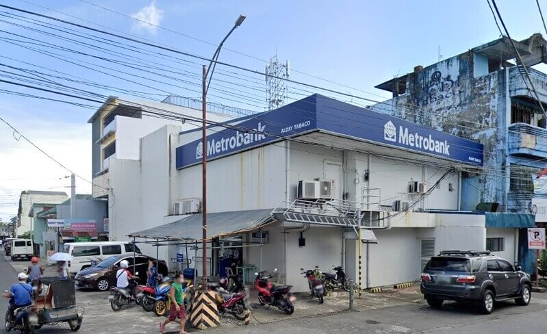A two-story Metrobank branch building located on a street corner in Albay-Tabaco. The building is white with a large blue wraparound sign featuring the white Metrobank logo and text