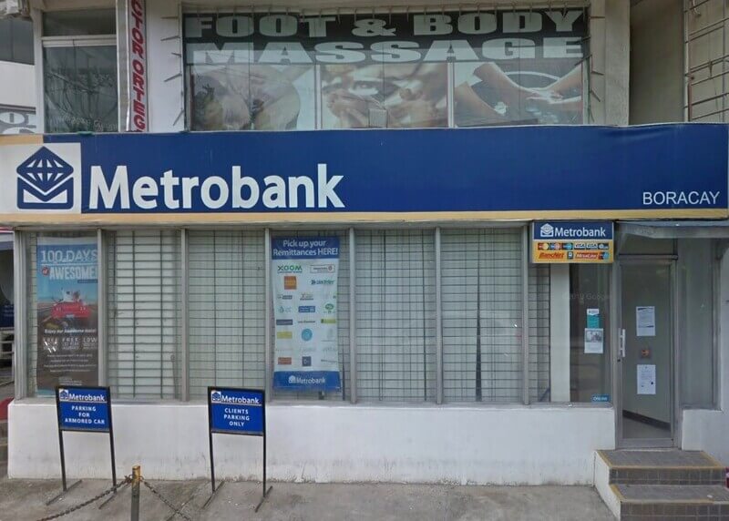 Exterior view of a Metrobank branch in Boracay, featuring a large blue sign with the bank's white logo. The storefront has glass windows with security bars, signage for remittance services, and designated parking signs for armored cars and clients. A "Foot & Body Massage" parlor is visible on the floor above