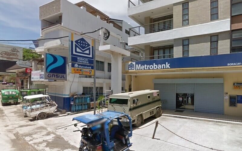 An armored truck and blue tricycles parked in front of the Metrobank Boracay - Station II branch building along a paved road. The bank features a large blue sign with the Metrobank logo, and a Petron Gasul signage is visible on the adjacent building