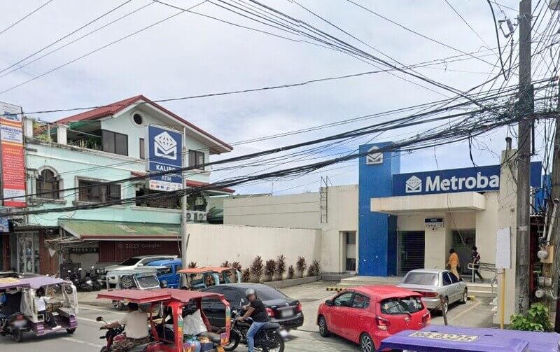 An exterior street-level view of the Metrobank Kalibo branch in the Philippines. The scene features a white and blue bank building with a prominent Metrobank sign and an ATM logo. In the foreground, there is a busy street with several motorized tricycles, a motorcycle, and parked cars under a network of overhead power lines