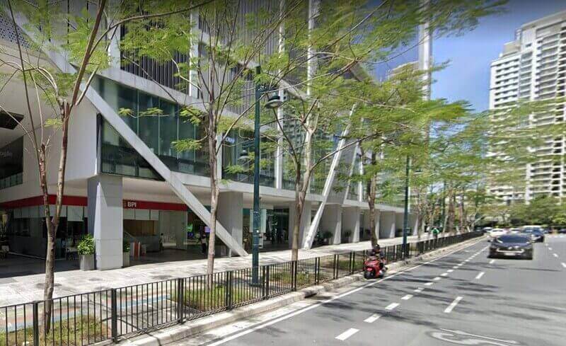 Exterior view of a BPI branch located at the ground floor of a modern white office building with diagonal structural beams on 26th Street, Bonifacio Global City (BGC), featuring a tree-lined sidewalk and a passing red scooter