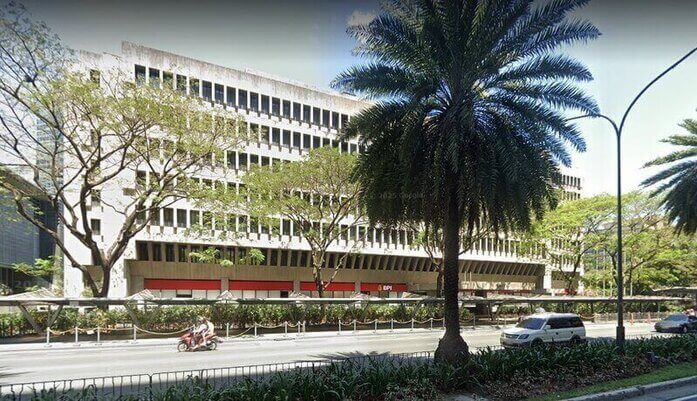 A wide exterior view of the SGV & Co. building on Ayala Avenue, featuring its distinct brutalist architectural style with vertical concrete fins, a red BPI branch at the ground level, and tropical trees lining the busy street in the foreground