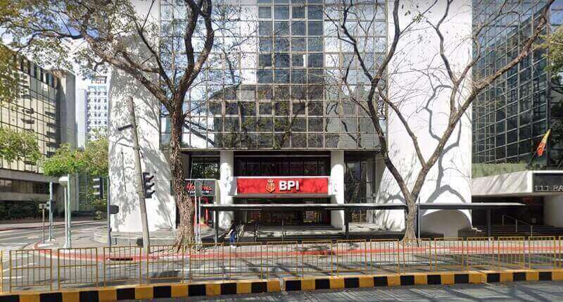 Exterior view of the BPI branch at Corinthian Plaza, showing the red building signage, glass facade, and sidewalk with yellow and black safety barriers