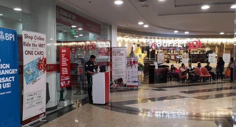 A BPI bank branch entrance located inside Glorietta Mall, featuring a security guard at a podium and promotional banners for debit cards. To the right, the entrance to Buffet 101 is visible with customers waiting in a seating area