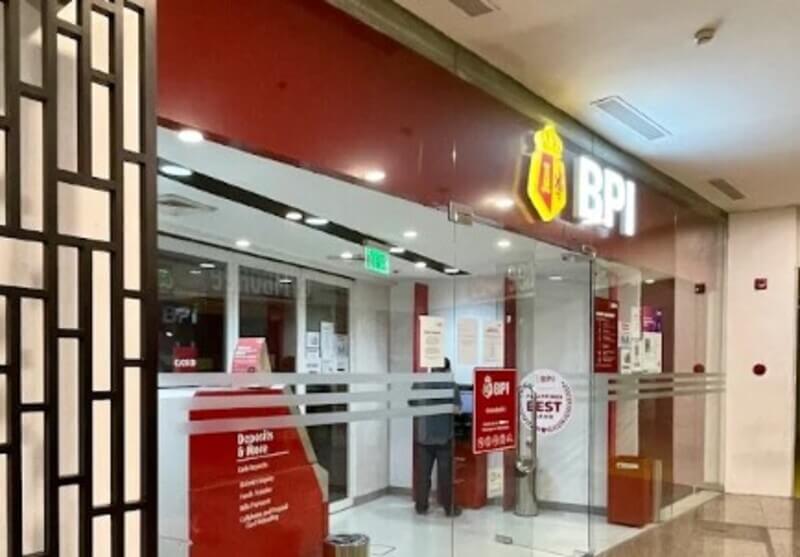Exterior view of a BPI (Bank of the Philippine Islands) branch located inside a mall, featuring glass doors, red and white branding, and a self-service kiosk near the entrance