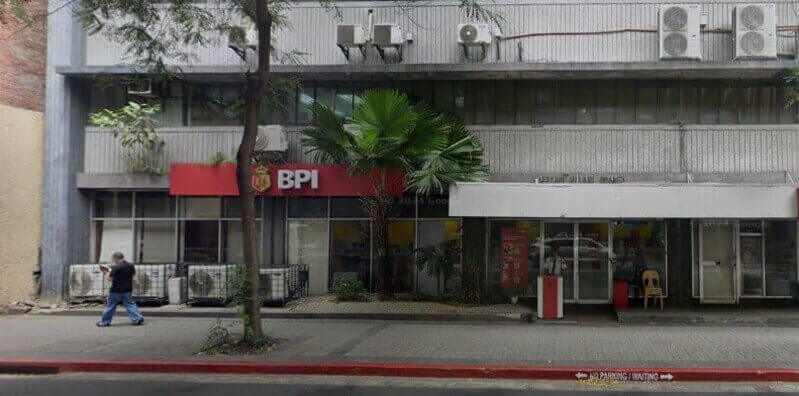 Exterior street-level view of a BPI (Bank of the Philippine Islands) branch featuring a red logo sign, glass windows, and a security desk at the entrance, with a pedestrian walking past on the sidewalk