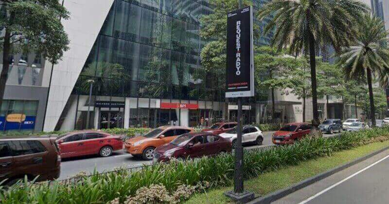 A street-level view of Ayala Avenue in Makati, Philippines, featuring the BPI branch at the base of a modern glass skyscraper. A vertical black banner in the foreground advertises the play "Request Sa Radyo" starring Lea Salonga and Dolly de Leon, with heavy traffic of colorful cars and tropical trees lining the sidewalk