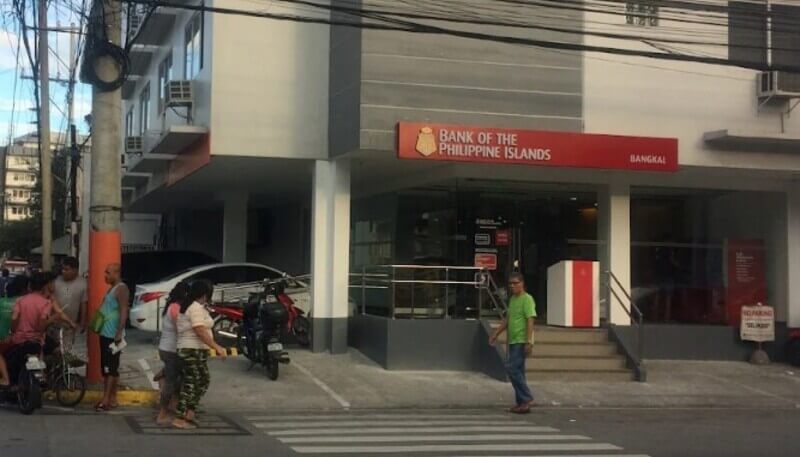 Exterior of a Bank of the Philippine Islands (BPI) branch in Bangkal, featuring a modern grey and white building with a prominent red and white logo sign above the entrance