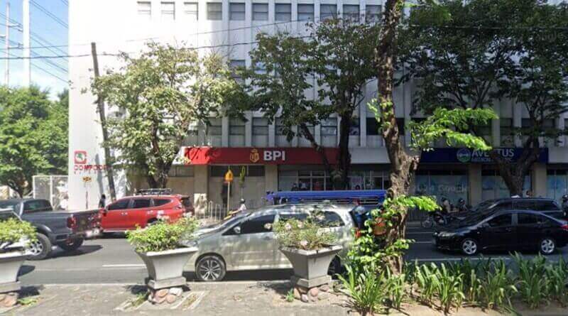Street view of a red BPI Buendia Pasong Tamo branch storefront located on the ground floor of a white multi-story building, with several cars and a blue jeepney passing by on a busy road lined with trees