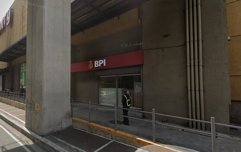 Exterior view of a BPI Cash And Carry Mall branch located at the ground level of Cash & Carry Mall. The image shows the red bank signage above a glass storefront, a security guard standing on the sidewalk, and a concrete support pillar in the foreground