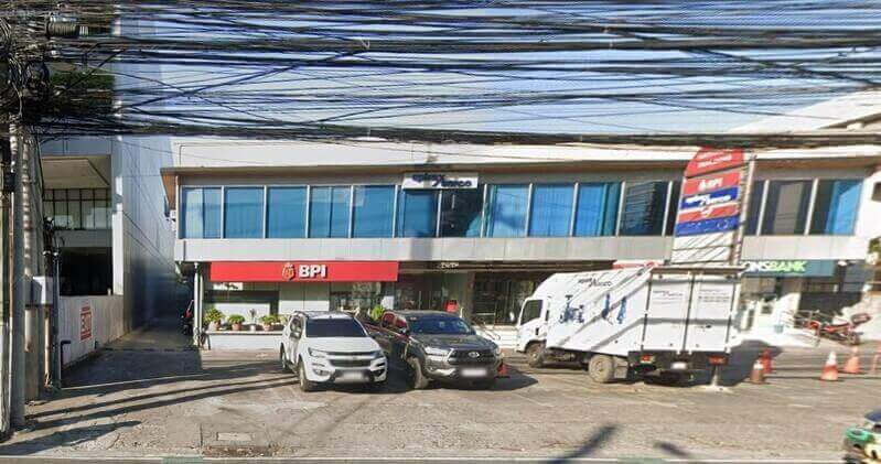 Street view of the BPI Chino Roces Ext branch office, a two-story building with a red BPI sign, glass windows, and several vehicles parked in front under a network of utility wires