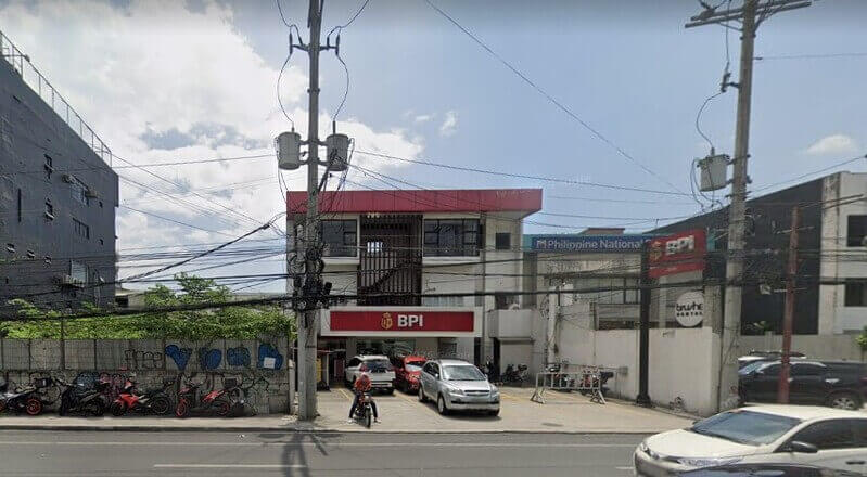 Exterior view of a red and white BPI (Bank of the Philippine Islands) branch located along a busy street with parked cars, motorcycles, and utility poles in the foreground