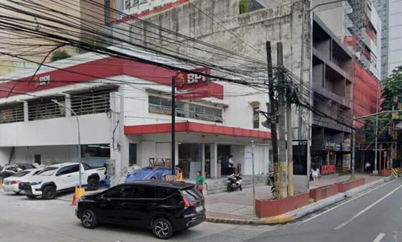 An external view of the BPI Espana-UST branch in Manila, featuring a two-story white building with a prominent red roofline and the BPI logo. Several vehicles, including a black SUV and a white pickup truck, are parked in front of the entrance, and a dense network of overhead power lines runs across the street