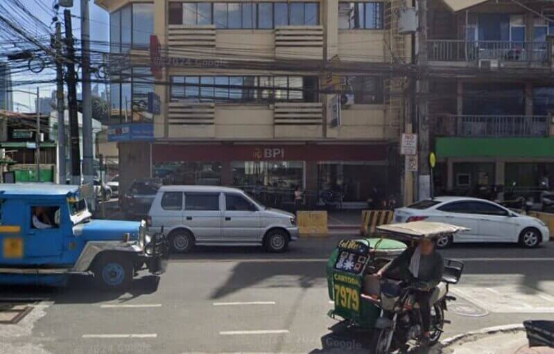A street-level view of a BPI JP Rizal - Binakod branch in the Philippines, featuring a blue jeepney and a green tricycle in the foreground with a multi-story commercial building behind them
