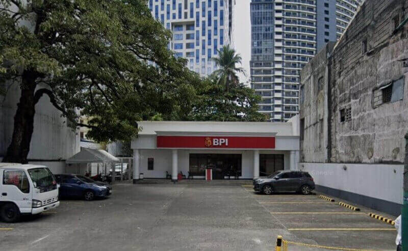 Exterior view of a white, single-story BPI branch with a prominent red sign, featuring a small front parking lot and framed by tall city buildings and a large tree