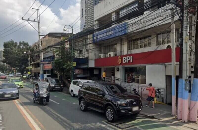 A street-level view of a BPI JP Rizal San Miguel branch with a prominent red sign, located in a multi-story building alongside a busy city road with parked cars, a black SUV, and a passing tricycle