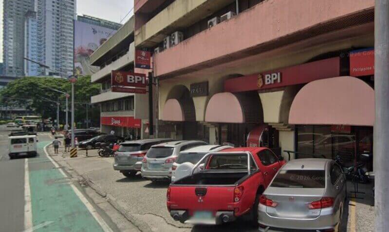 Street-level view of a BPI branch located along a busy road (EDSA) with several cars parked in front. The building features red signage and pink arched awnings, with a PSBank branch visible in the background and city skyscrapers in the distance