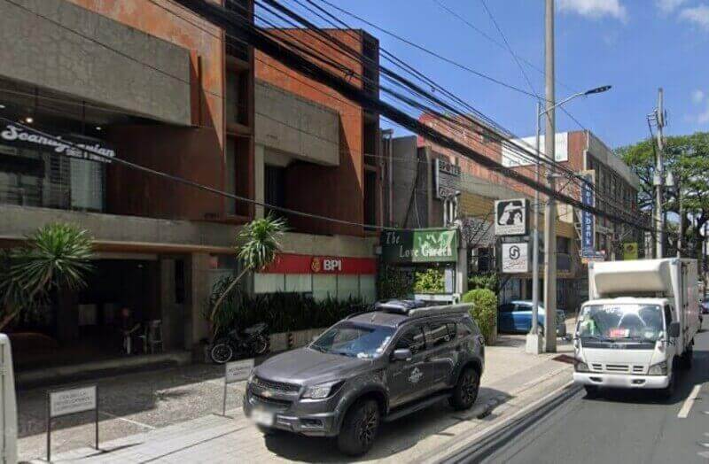 A street-level view of a BPI branch on Pasay Road, featuring a modern brown and concrete building with a red BPI sign, a gray Chevrolet SUV parked in front, and a white delivery truck driving on the road
