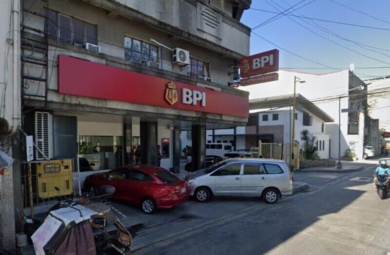 Exterior view of a BPI San Antonio Kamagong branch located at the corner of a city street, featuring its signature red signage, an outdoor ATM, and cars parked in front of the building
