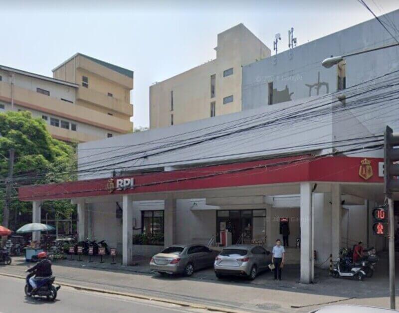 Exterior of a BPI Tayuman branch featuring a wide red awning, white pillars, and a parking area with cars and motorcycles in front of a multi-story building