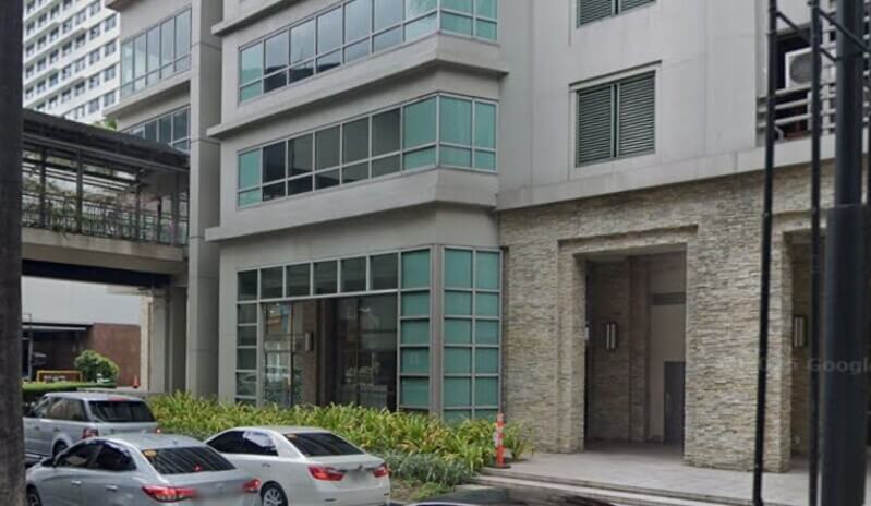Exterior view of the BPI branch at The Residences at Greenbelt, featuring a modern grey multi-story building with large glass windows, a stone-cladded entrance, and cars parked along the street in the foreground