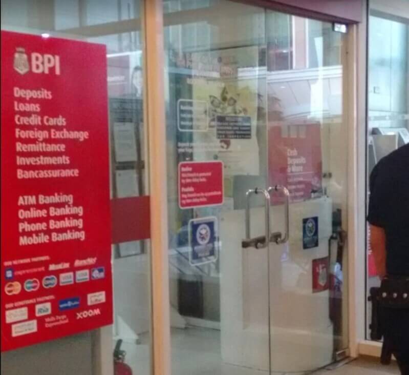 The entrance to a BPI (Bank of the Philippine Islands) branch, featuring glass double doors and a prominent red sign listing services such as Deposits, Loans, and Foreign Exchange. The sign also displays various partner logos including Mastercard, Visa, and Xoom