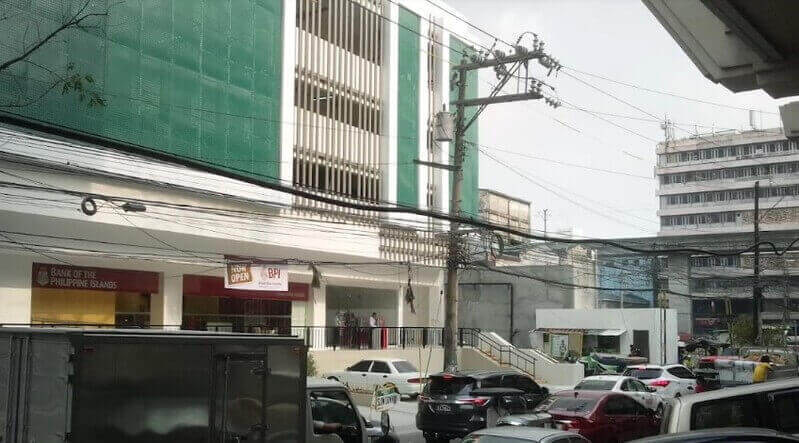 Exterior view of the Bank of the Philippine Islands (BPI) branch at Amaia Skies Avenida, featuring a multi-story building with green safety netting on the upper floors and a street-level facade with red BPI signage overlooking a busy urban road with traffic